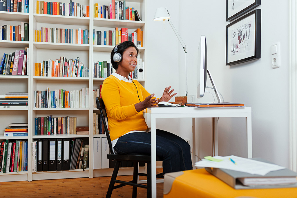 Frau sitzt im Wohnzimmer mit Headset vor ihrem Computer und nimmt an einer Videokonferenz teil