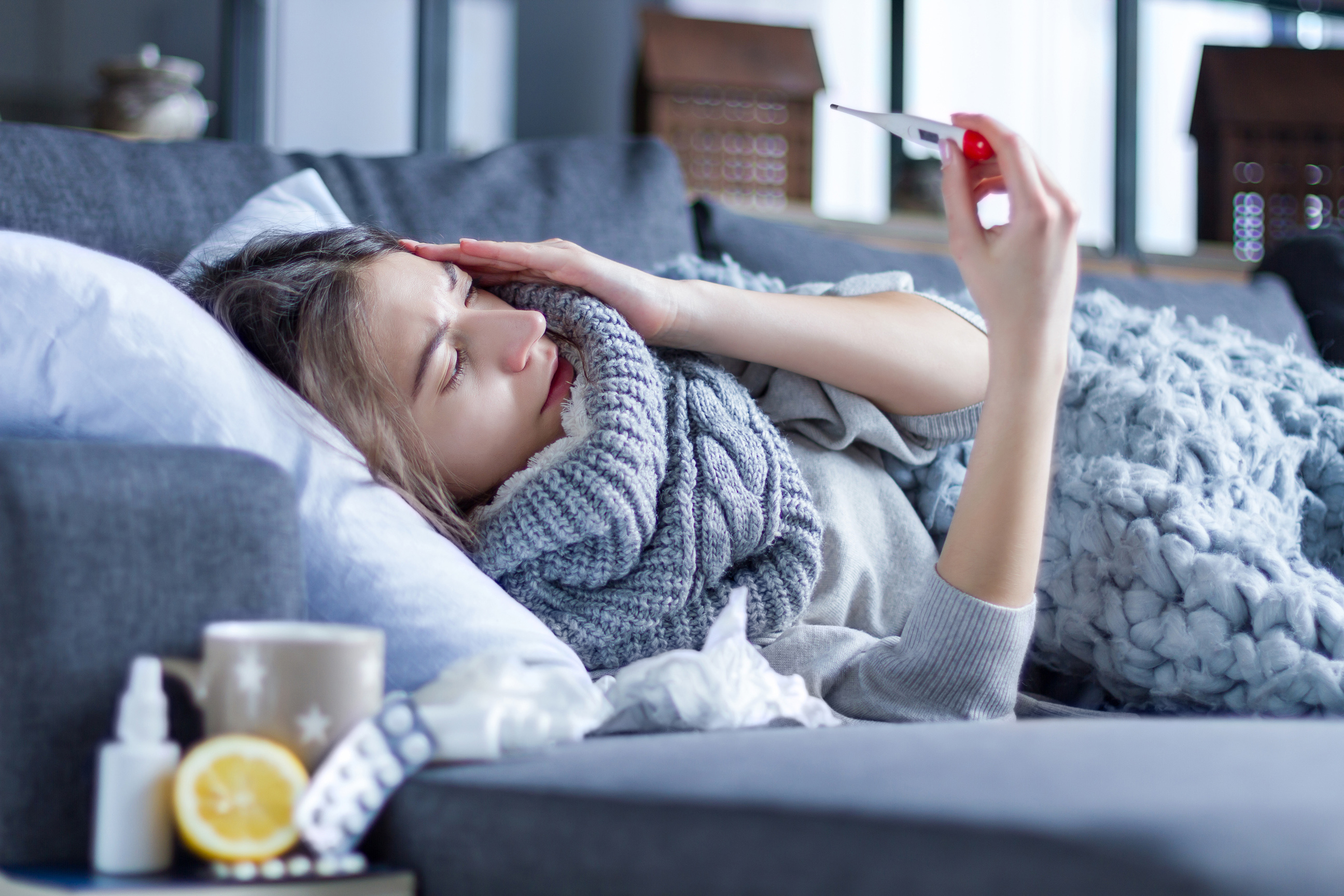 Kranke junge Frau mit Schal liegt in eine Decke gehüllt im Bett und misst die Temperatur mit einem Thermometer. 