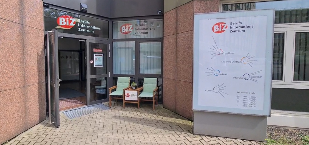 Blick auf den Eingang des BiZ: Neben einem Infoplakat befindet sich eine Sitzbank, daneben ist die geöffnete Tür zu sehen