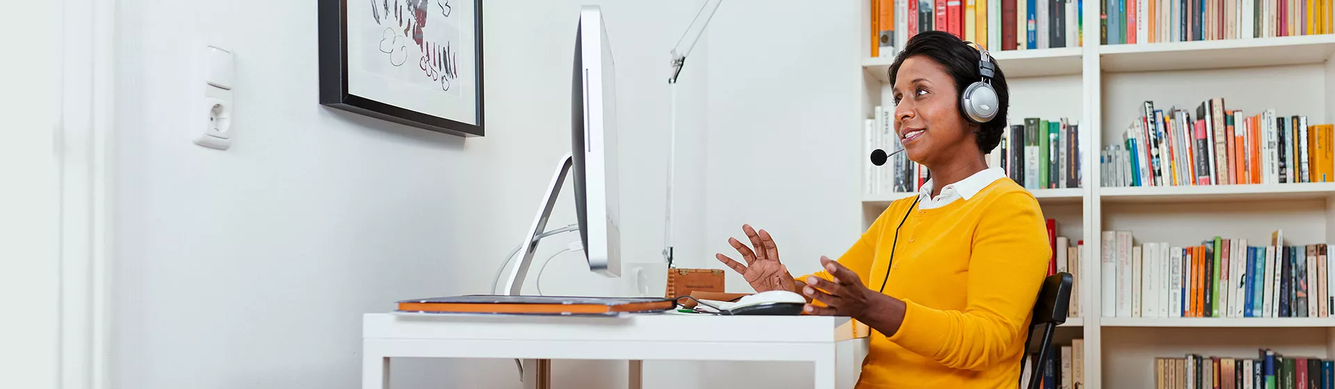 Frau sitzt im Wohnzimmer mit Headset vor ihrem Computer und nimmt an einer Videokonferenz teil