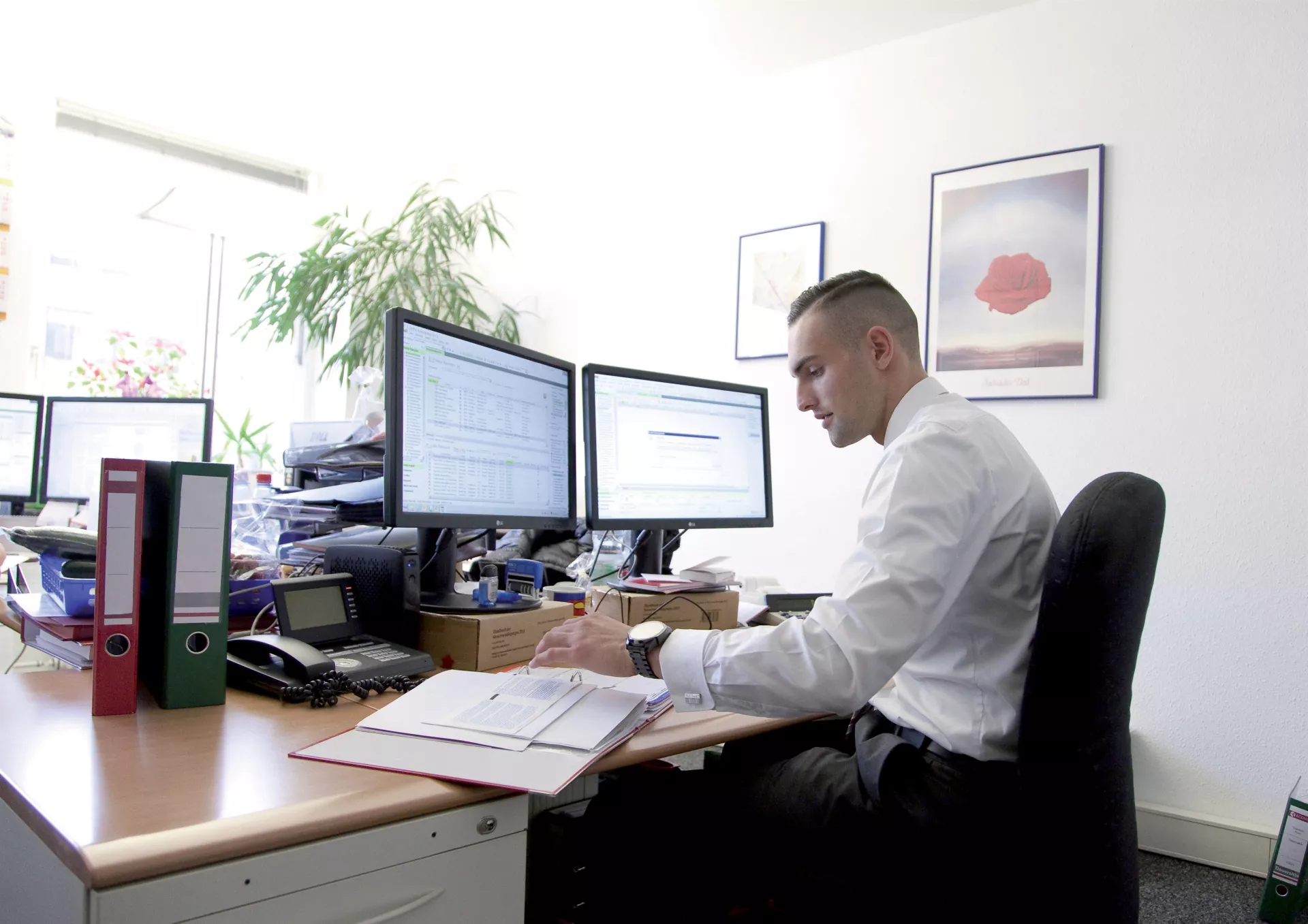 Ein junger Mann sitzt an einem Schreibtisch vor zwei PC-Monitoren und schaut auf einen Aktenordner.