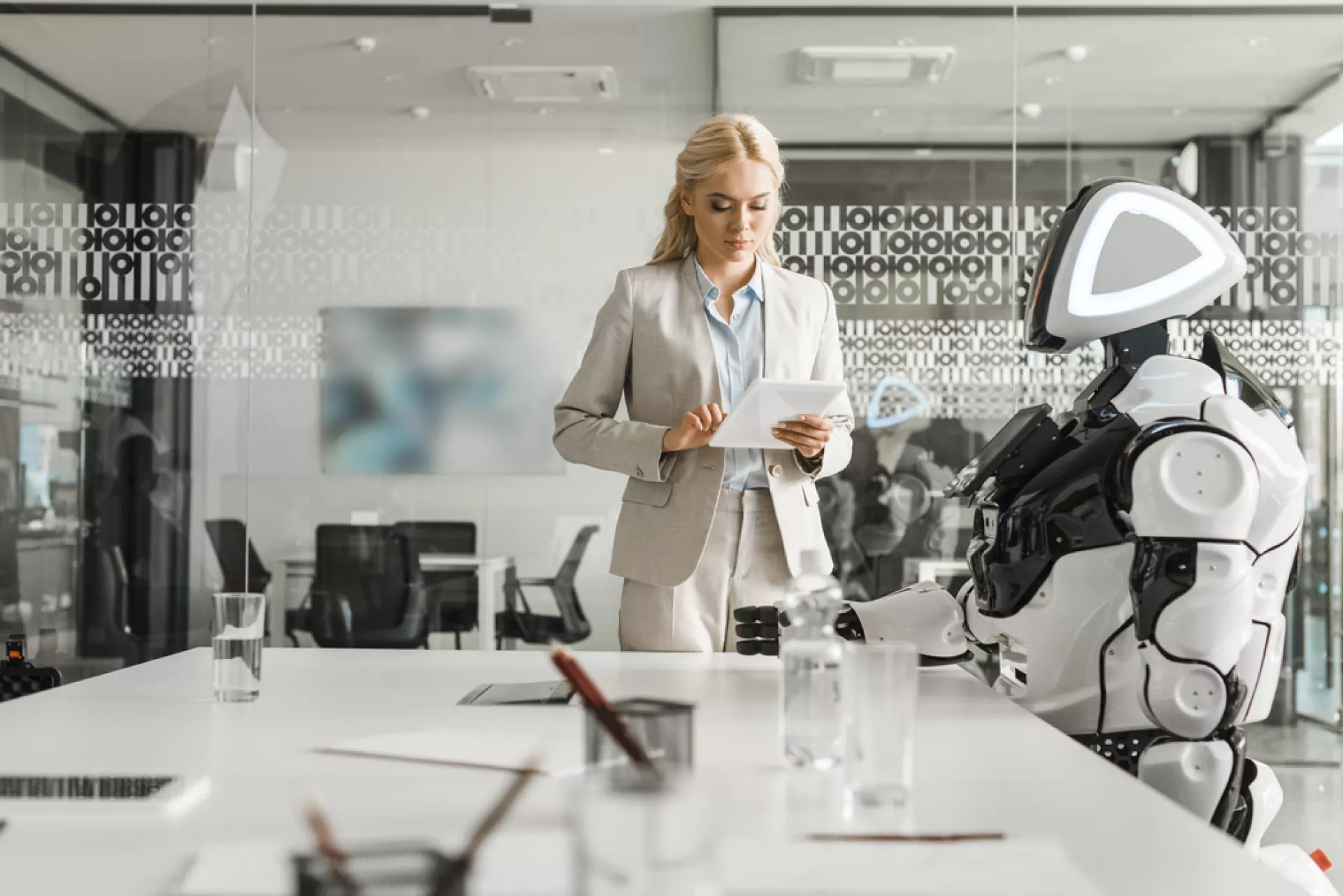 Auf dem Foto steht eine Frau in Businesskleidung in einer modernen Bürolandschaft vor einem Besprechungstisch, an dem ein Roboter sitzt.