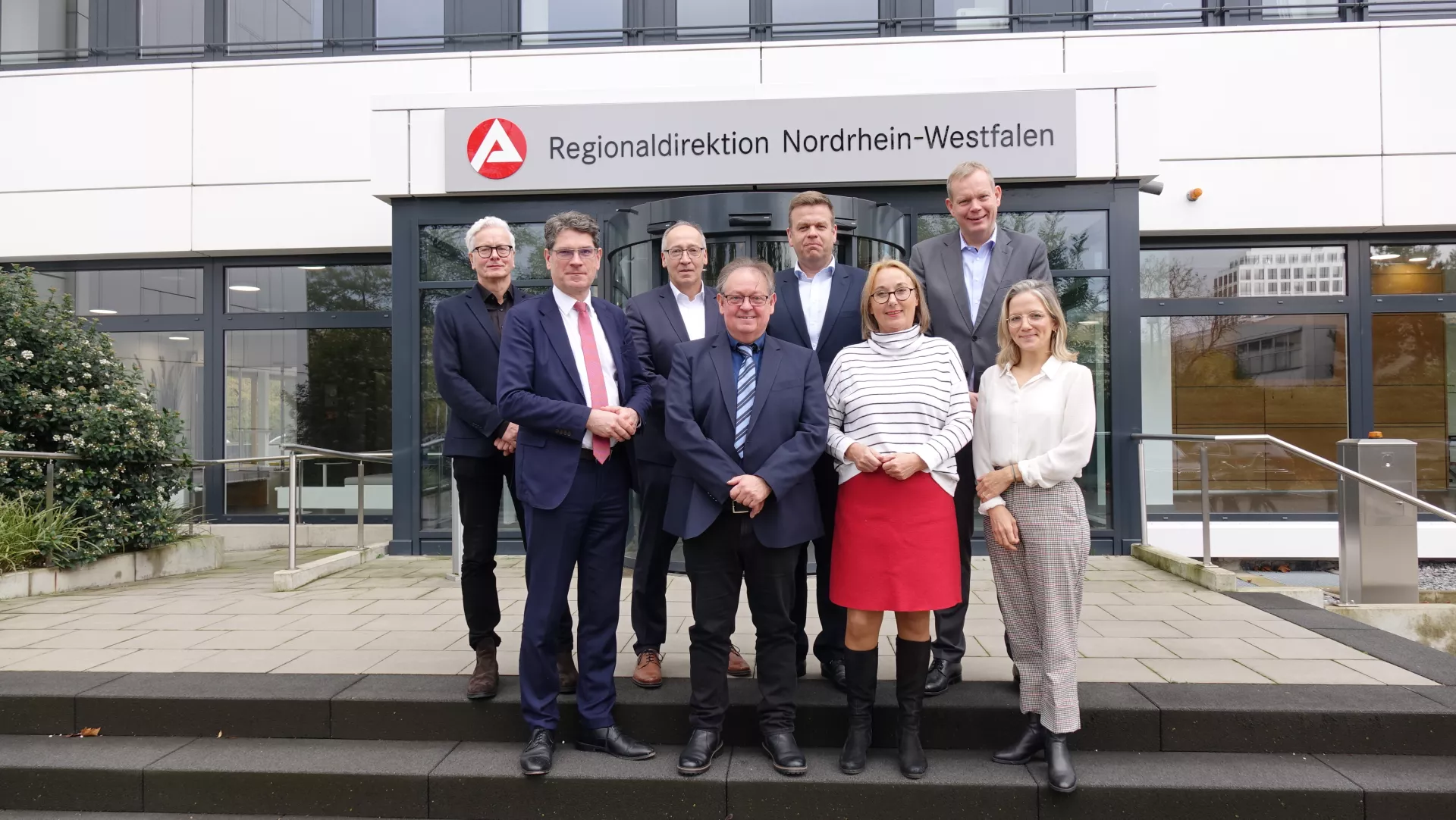 Auf dem Bild sieht man die Mitglieder des arbeitsmarktpolitischen Beirates der Regionaldirektion NRW vor dem Gebäude der Regionaldirektion.  Im Hintergrund  ist das Logo der Regionaldirektion über dem Hauseingang zu sehen. 