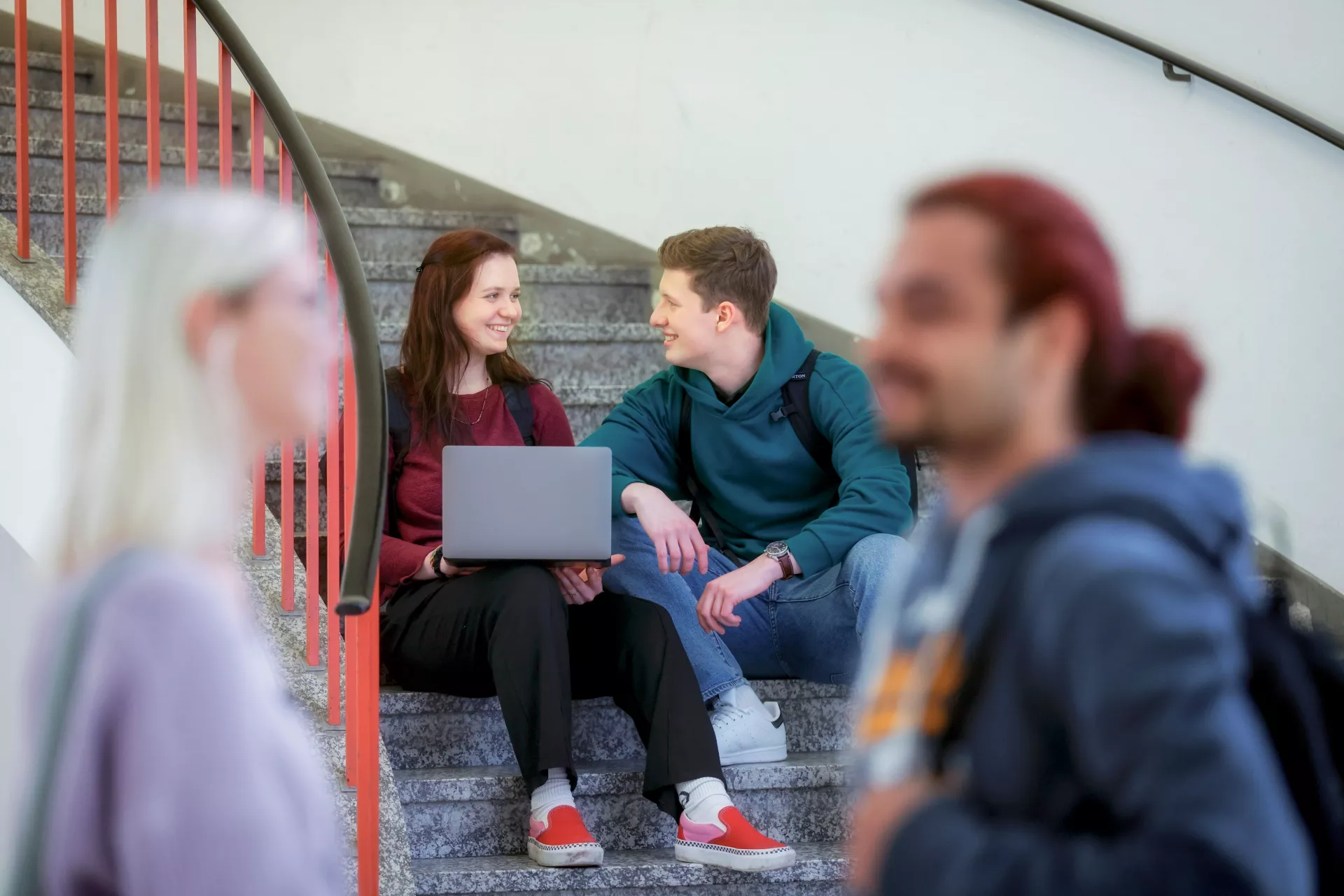 Eine Schülerin und ein Schüler sitzen auf einer Treppe eines Universitätsgebäude und schauen auf einen Computer