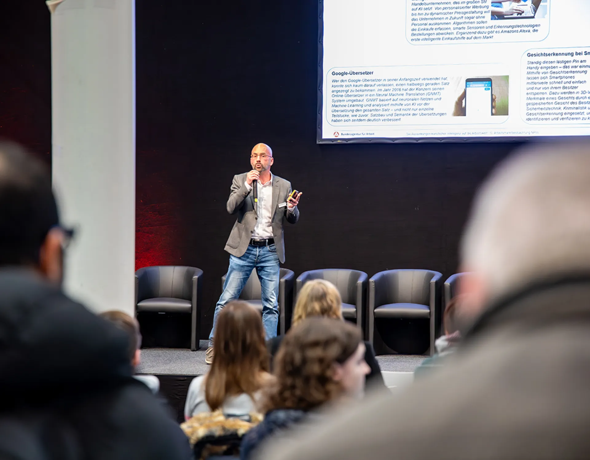 Ein Mann referiert auf einer Bühne. Im Vordergrund sitzen Zuhörer.