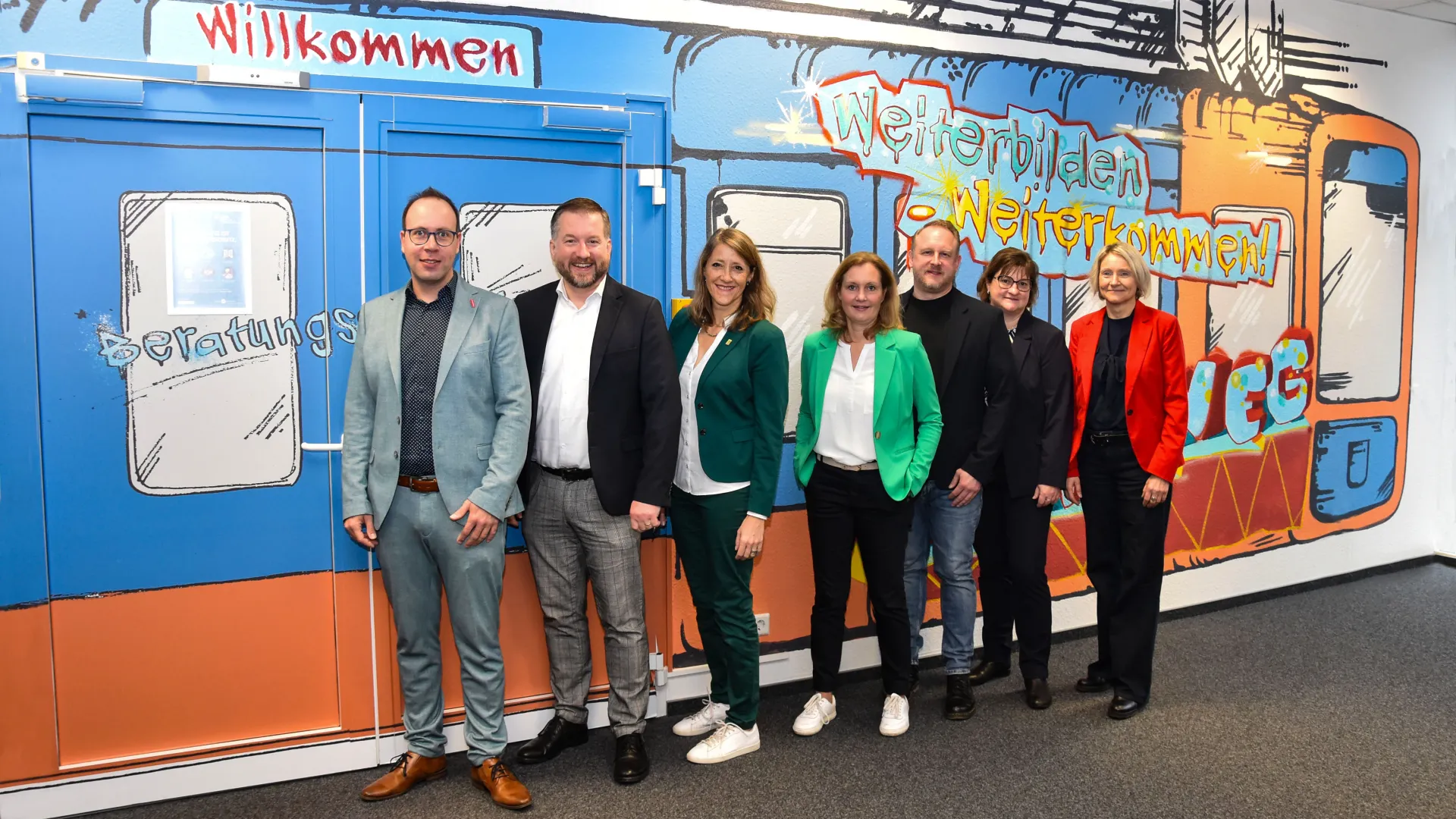 Die sieben Marktpartner im Bergischen Städtedreieck vor einer orange-blauen Zeichnung der Schwebebahn. Vier Damen und drei Herren stehen in einer Reihe.