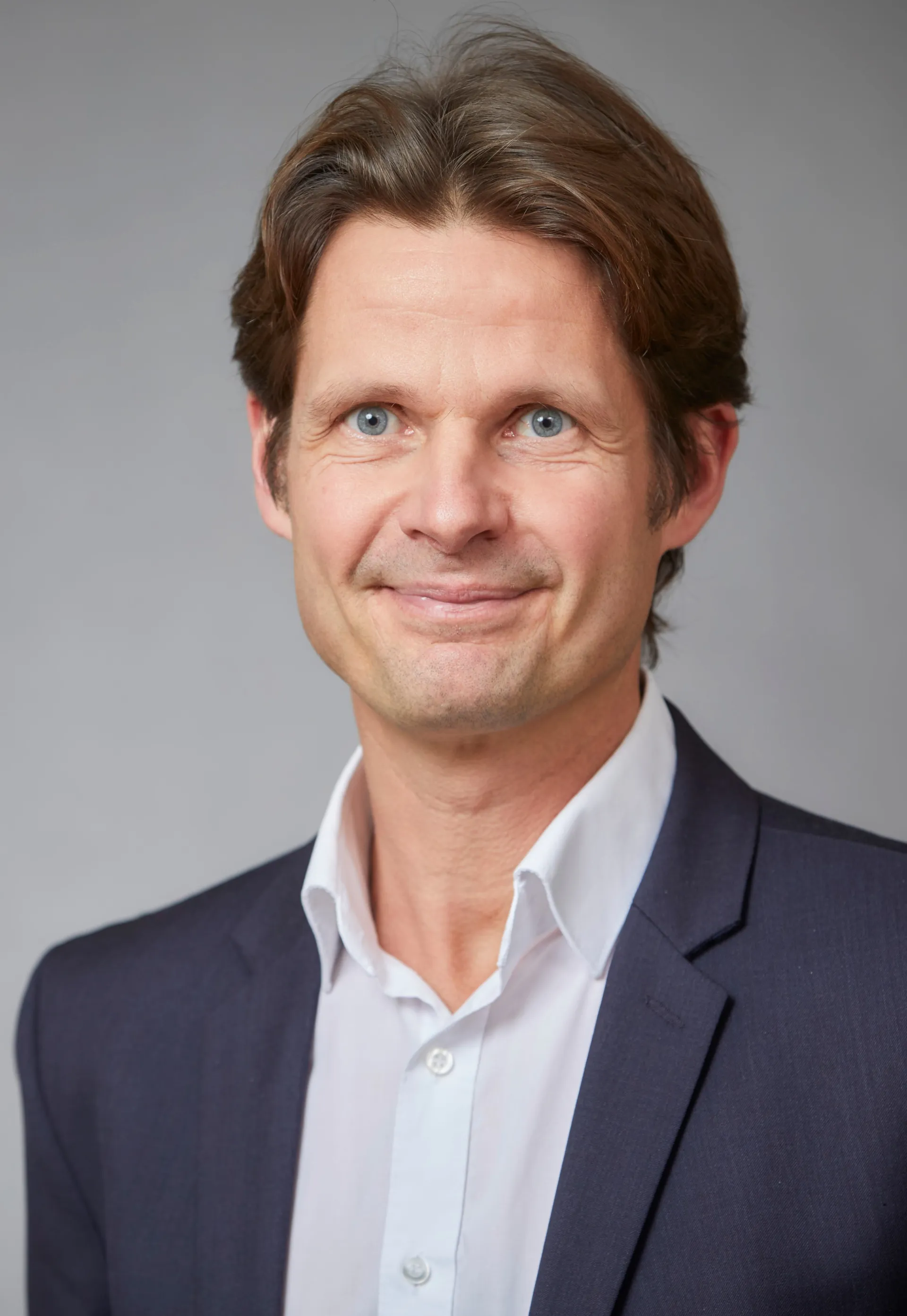 Ein Mann in einer dunkelblauen Anzugjacke und in einem weißen Hemd lächelt in die Kamera.
