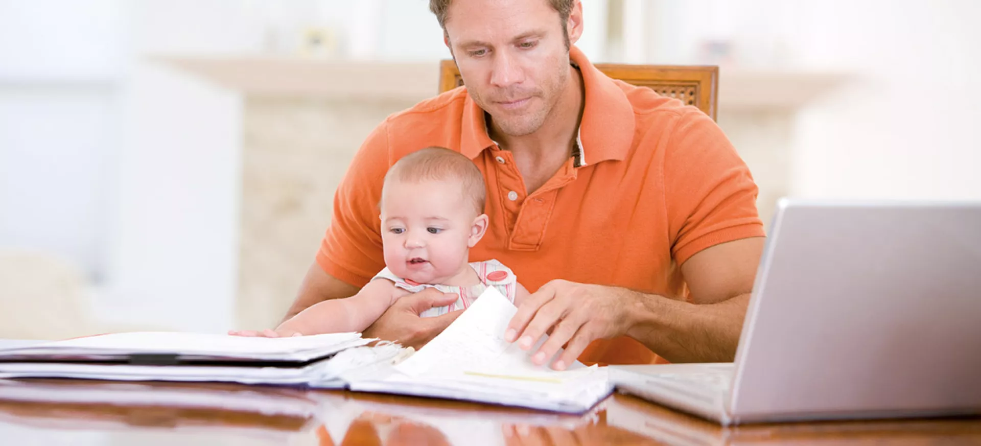 Mann mit Baby auf dem Schoß sieht Akten und Broschüren durch um einen Antrag auf finanzielle Unterstützung zu stellen.