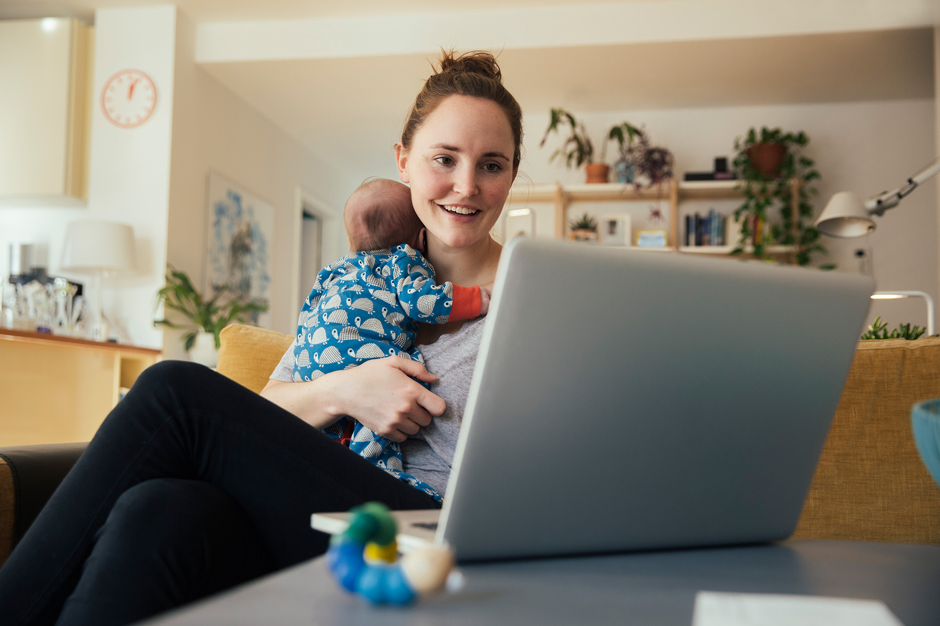 Frau mit Baby auf dem Arm schaut zuhause auf den Bildschirm eines Laptops