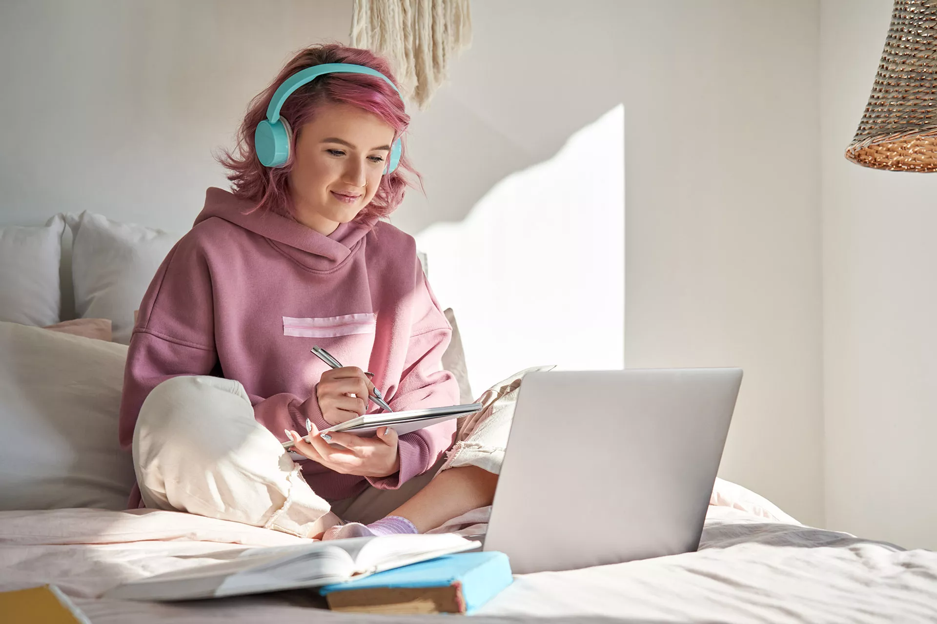 Mädchen mit rosa Haaren und Kopfhörern sitzt auf ihrem Bett und arbeitet mit Block und Laptop