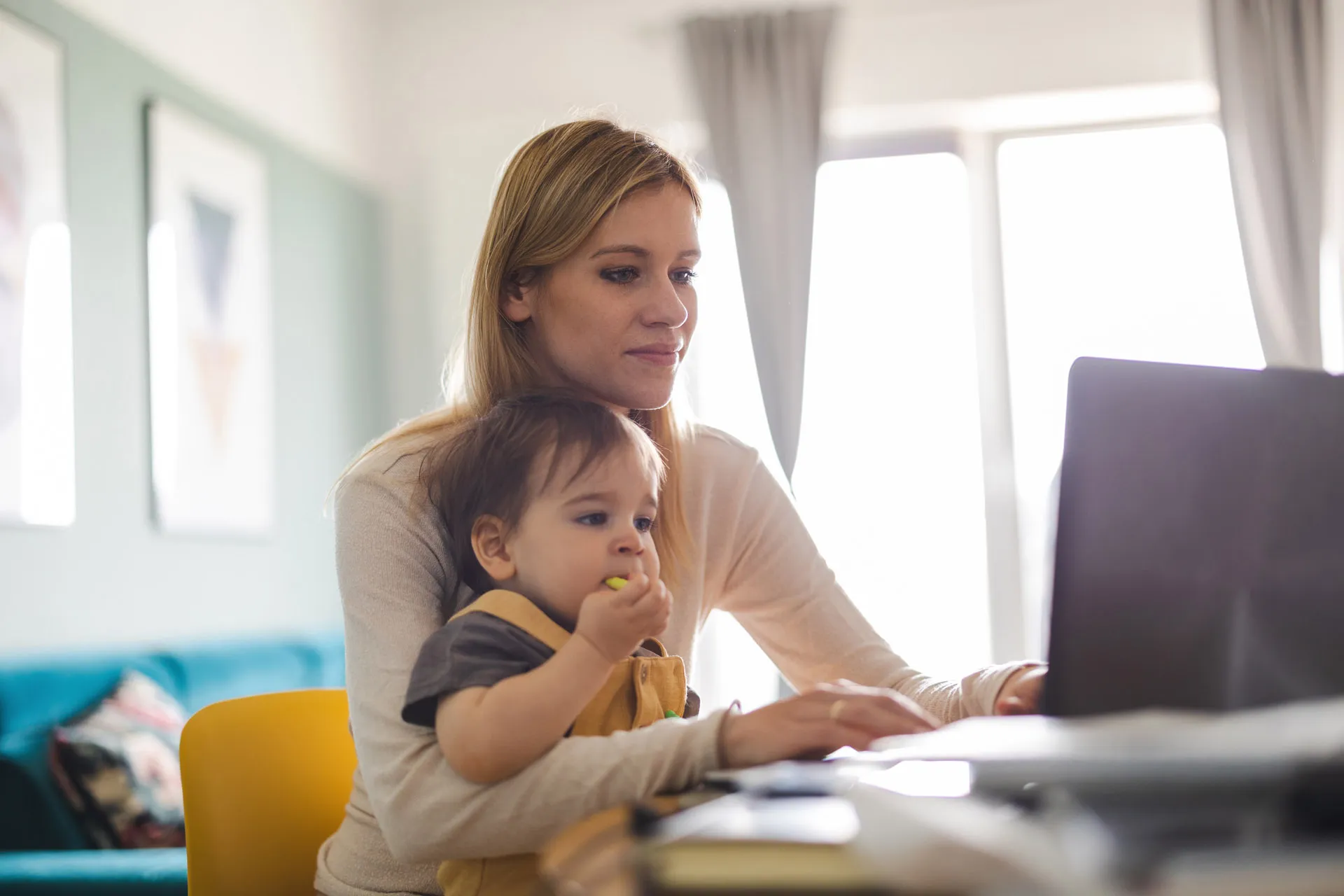 Eine junge Frau schaut auf einen laptop-Bildschirm und Ihr Kleinkind sitzt neben ihr.