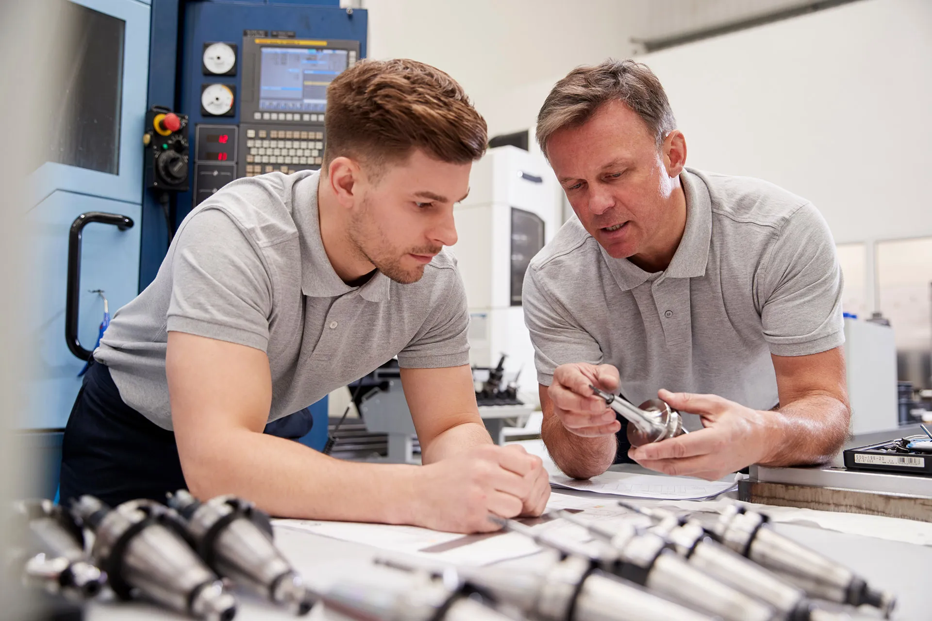 Ein Ingenieur zeigt einem jungen Auszubildenden in einer Werkstatt, wie man CAD-Zeichnungen bemisst.