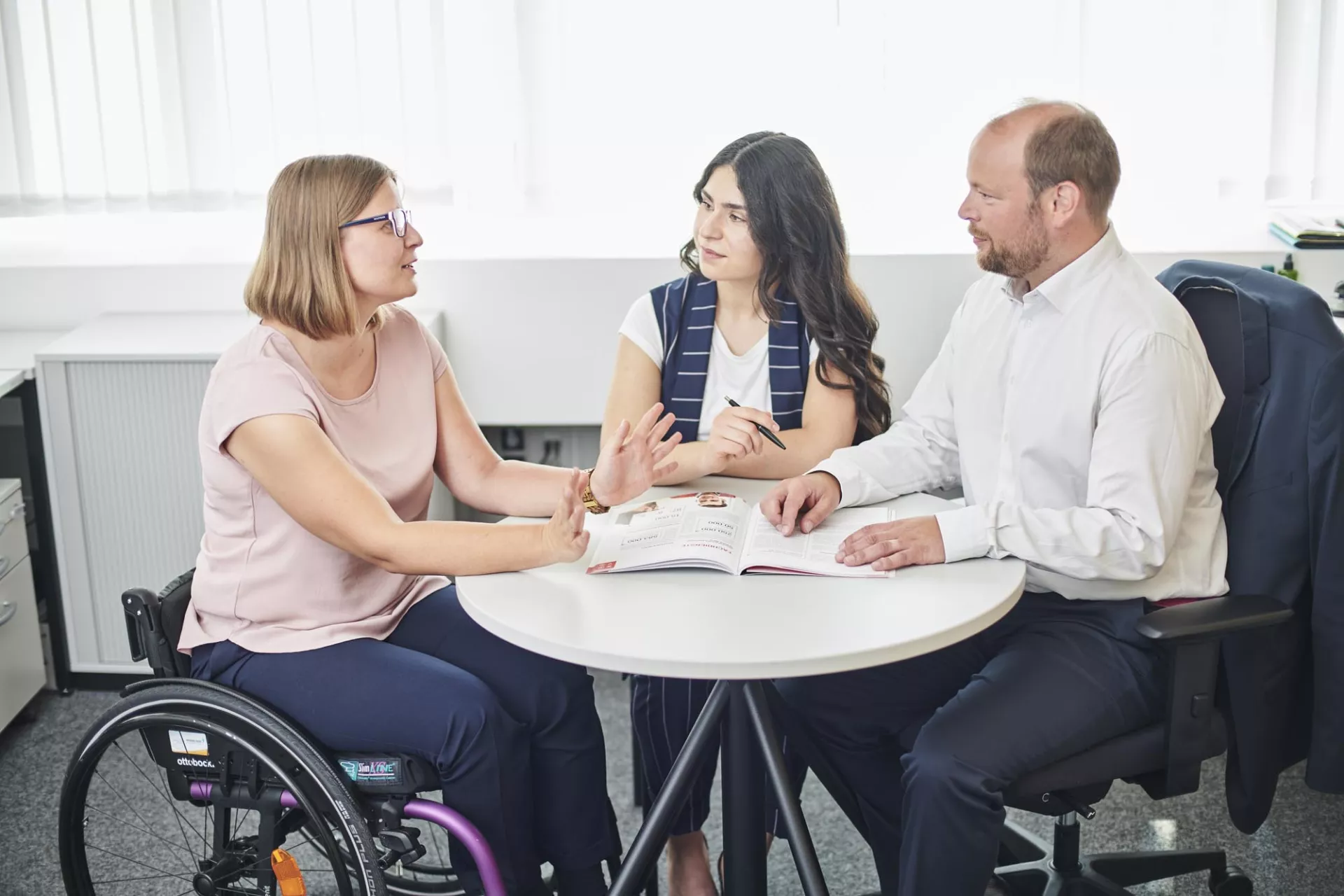 Drei Personen sitzen um einen Tisch und reden. Eine davon sitzt in einem Rollstuhl.