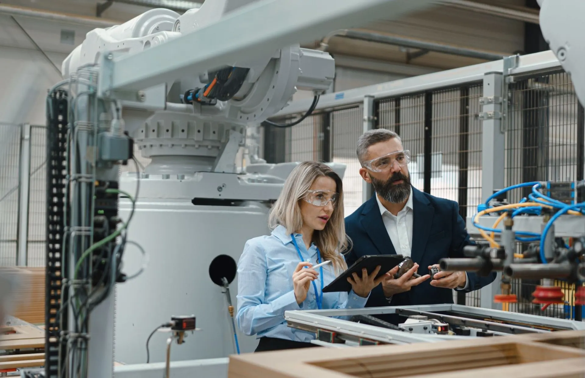 Eine Ingenieurin und ein Projektmanager stehen in einer modernen Industriehalle neben einem Präzisionsroboterarm. 